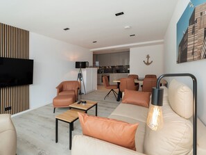 Ein modernes Wohnzimmer mit einer Couch, Sesseln, einem Couchtisch und einem Fernseher. Es gibt einen Essbereich mit Tisch und Stühlen, und im Hintergrund eine Küche.