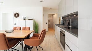 Cuina moderna amb armaris blancs, tauleig negre i electrodomèstics d'acer inoxidable. Hi ha una zona de menjador amb taula de fusta i cadires de pell.