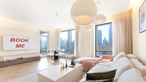 A modern living room with a large flat-screen TV, a sofa, a coffee table, and a city view through large windows. - Stayshort - Luxury 2 Bedroom in Downtown That Sleeps 4 With Balcony (Dubai)