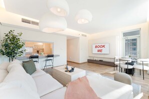 A modern living room with a sofa, coffee table, and a dining area with chairs and a table. A sign that reads "BOOK ME" is visible on the wall. - Stayshort - Luxury 2 Bedroom in Downtown That Sleeps 4 With Balcony (Dubai)