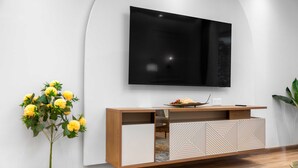 A modern living room with a flat-screen TV mounted on a white wall, a wooden console with geometric patterns, and a potted plant with yellow flowers. - Stayshort - High-floor 1 Bedroom in Dubai Marina That Sleeps 2 (Dubai)