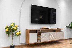 Ein modernes Wohnzimmer mit einem an einer weißen Wand montierten Flachbildfernseher, einer Holzkonsole mit geometrischen Mustern und einem Blumentopf mit gelben Blüten.
