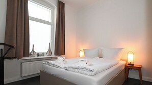 Un lit bien fait, avec du linge de lit et des serviettes blanches, une table de chevet avec une lampe et une fenêtre avec des rideaux.