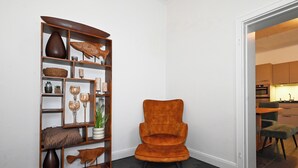 Un coin aménagé avec une étagère en bois où sont exposés divers objets décoratifs, un fauteuil moelleux de couleur orange et une porte-fenêtre donnant sur un espace cuisine.