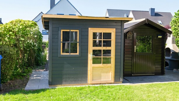 Una piccola struttura di legno verde scuro con una finestra e una porta, accanto a un edificio più grande con tetto spiovente.