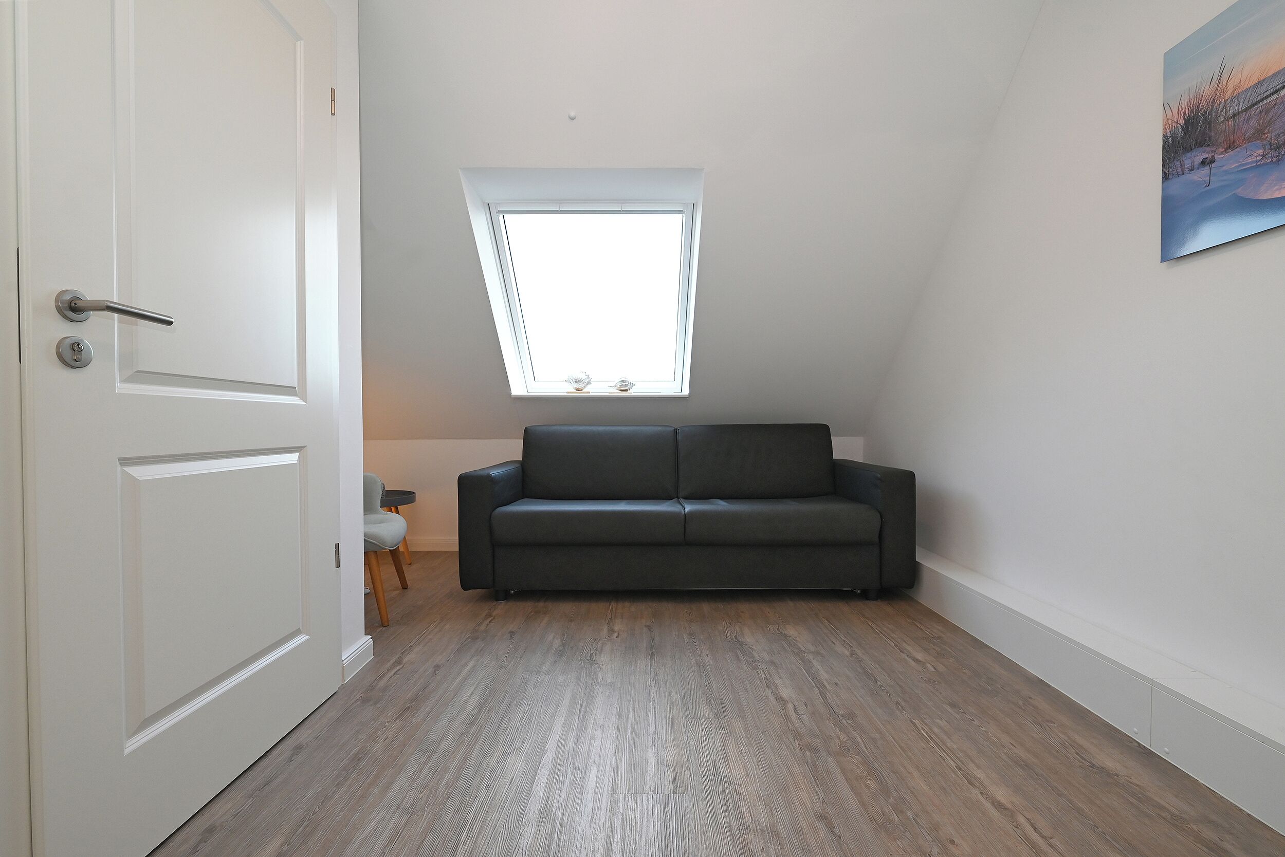 A room with a sloped ceiling, a window, a dark-colored sofa, and a wooden floor.