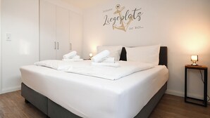 Un letto rifatto con lenzuola e asciugamani bianchi, una testiera scura e un muro con un'insegna decorativa a forma di ancora.