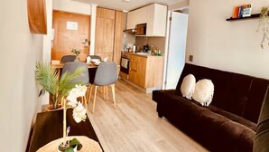 Ein modernes Wohnzimmer mit einer dunkelbraunen Couch, einem runden gewebten Tisch und einem Essbereich mit Stühlen und einer kleinen Pflanze.