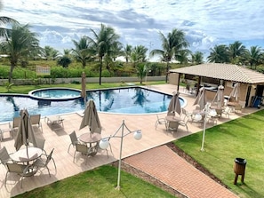 A swimming pool with lounge chairs and umbrellas, surrounded by palm trees and a covered seating area. - Beachfront Paradise in Condo w/ Pool in Itacimirim - Rsvs (Camaçari)