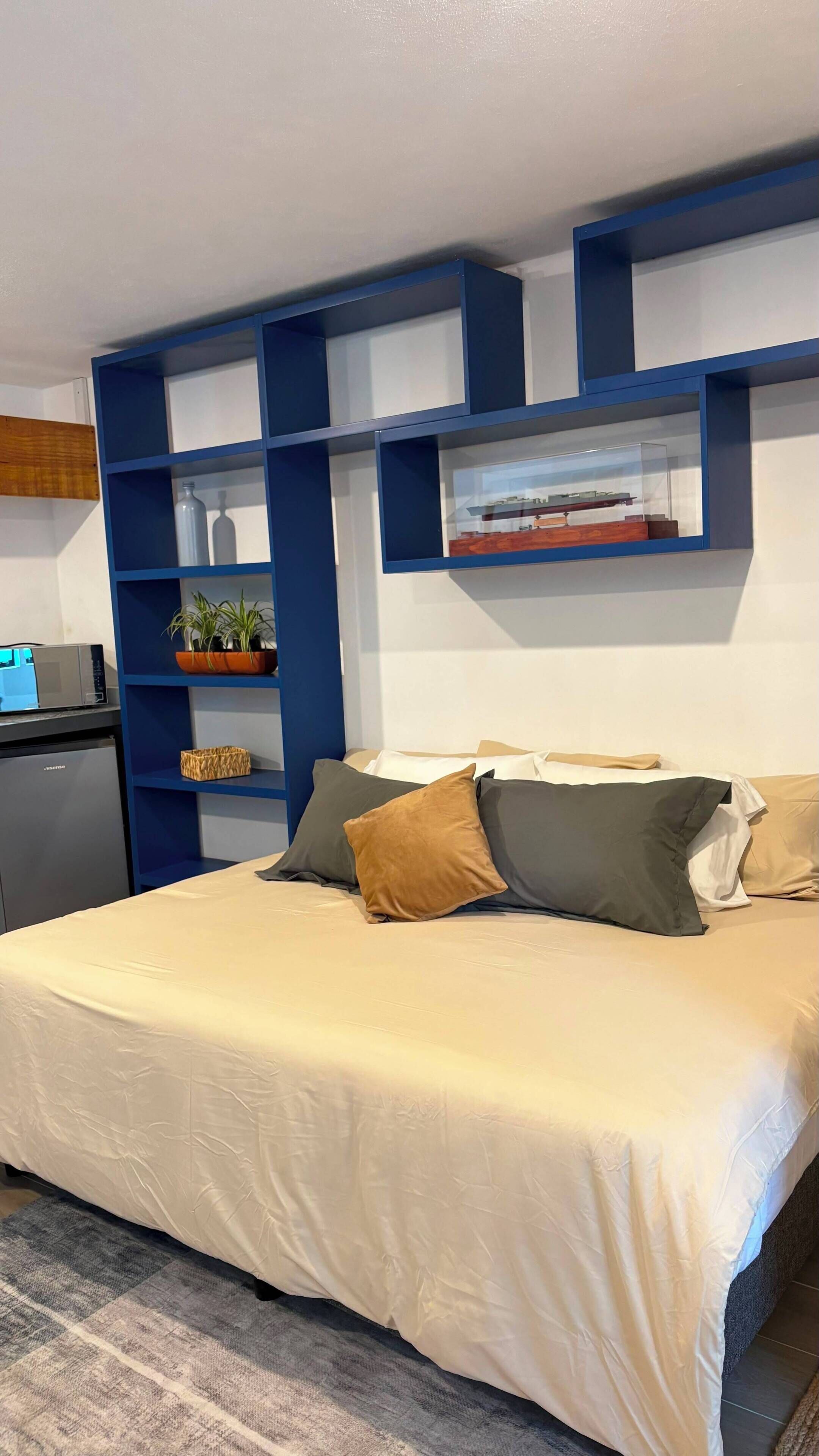 Un dormitorio con una cama, almohadas, un escritorio con una computadora y una repisa con objetos decorativos.