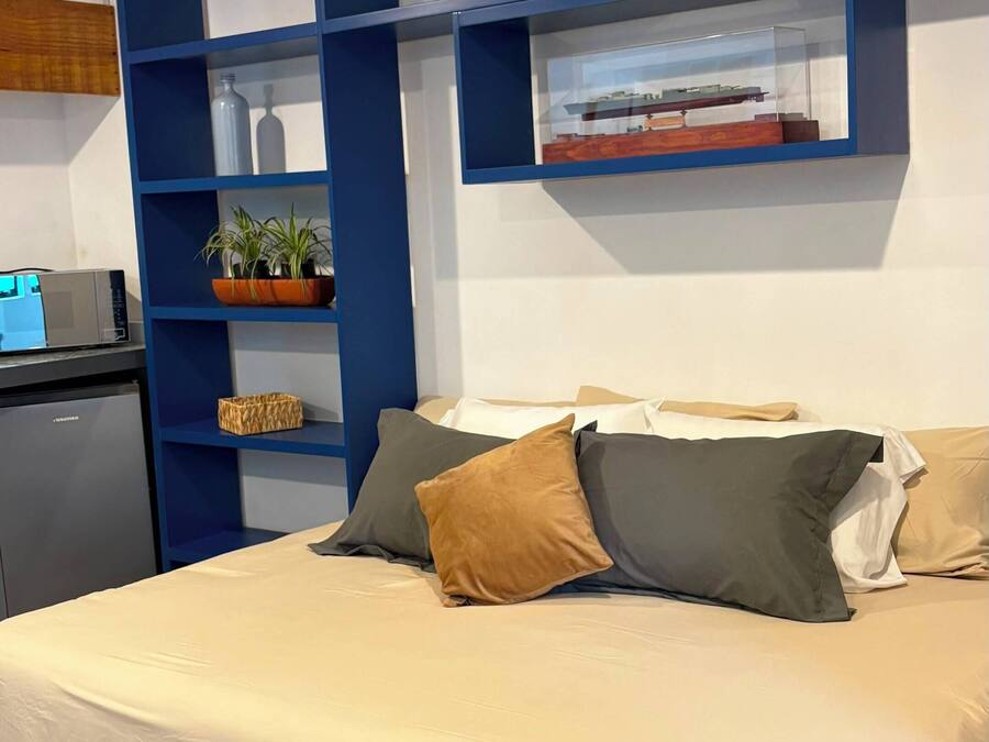 Un dormitorio con una cama, almohadas, un escritorio con una computadora y una repisa con objetos decorativos.