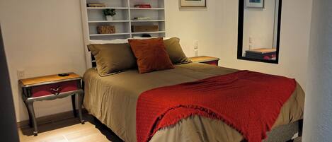 A bedroom with a bed, a nightstand, a dresser, a shelf with decorative items, and a mirror.