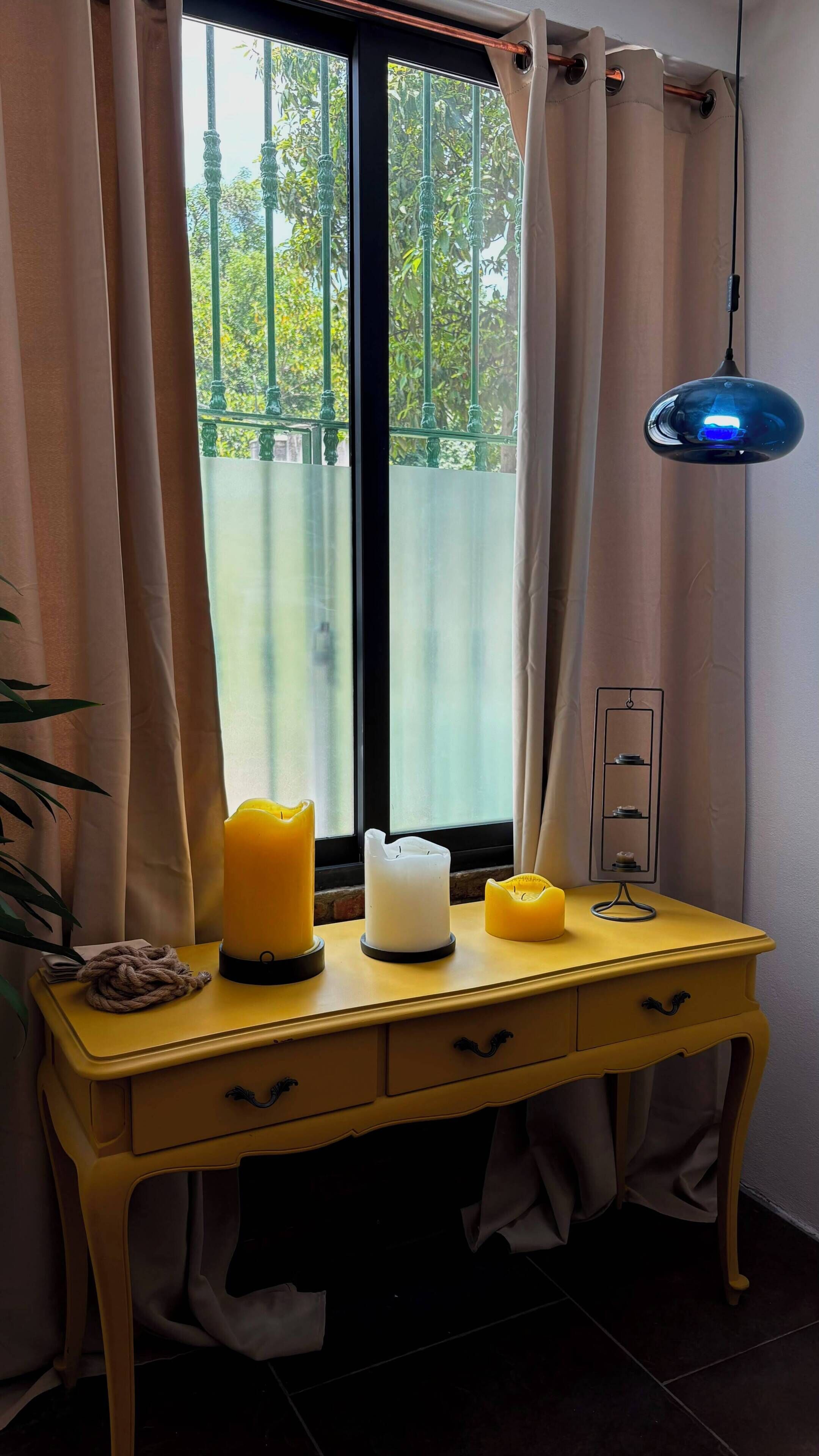 Una habitación con un escritorio amarillo, una ventana con cortinas, una lámpara colgante azul y una vela.