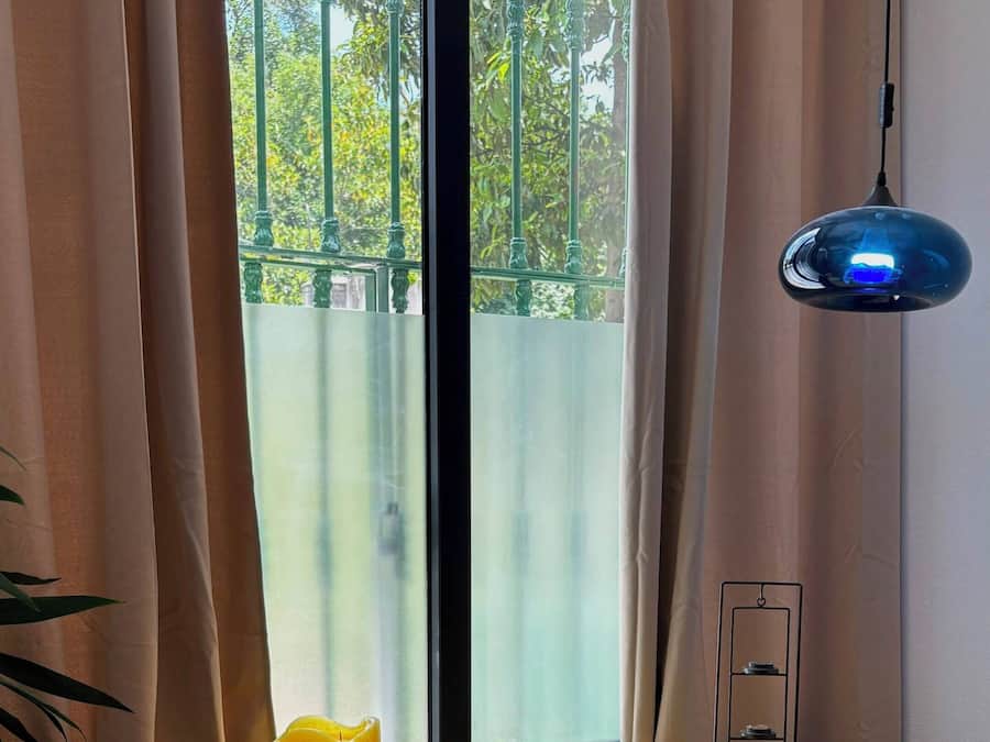 Una habitación con un escritorio amarillo, una ventana con cortinas, una lámpara colgante azul y una vela.