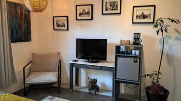 Une chambre d’hôtel avec un lit, un bureau avec une télévision, une chaise, un petit meuble, une plante en pot et des œuvres d’art accrochées au mur.