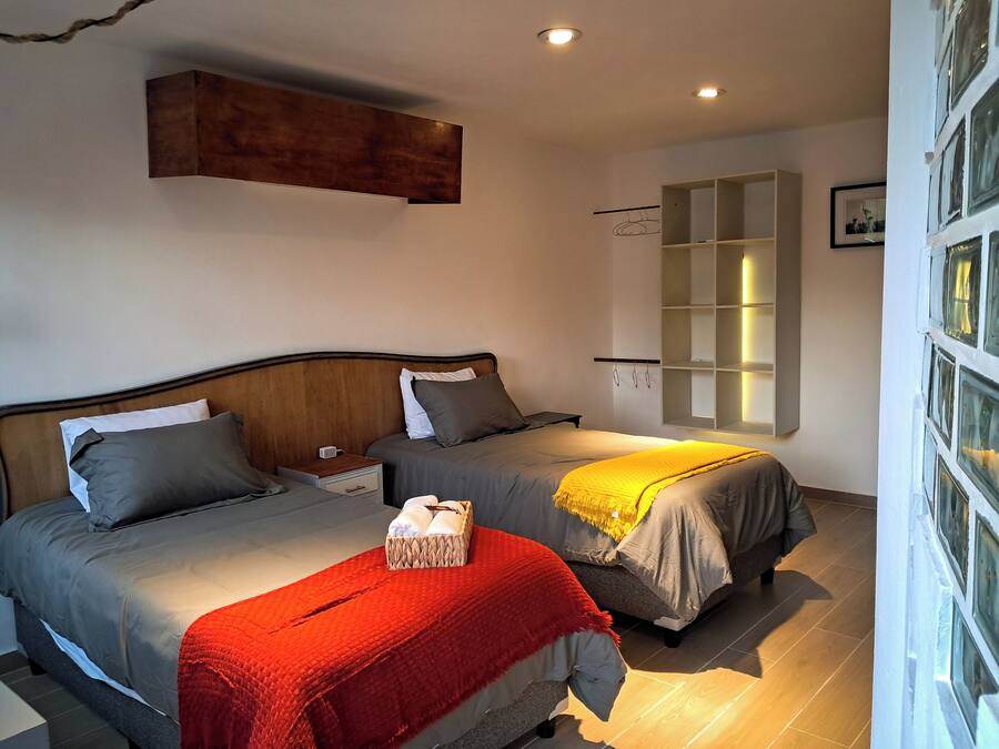 Een slaapkamer met twee bedden, een nachtkastje, een rode deken, een houten hoofdbord, een wandplank en een raam met gordijnen.