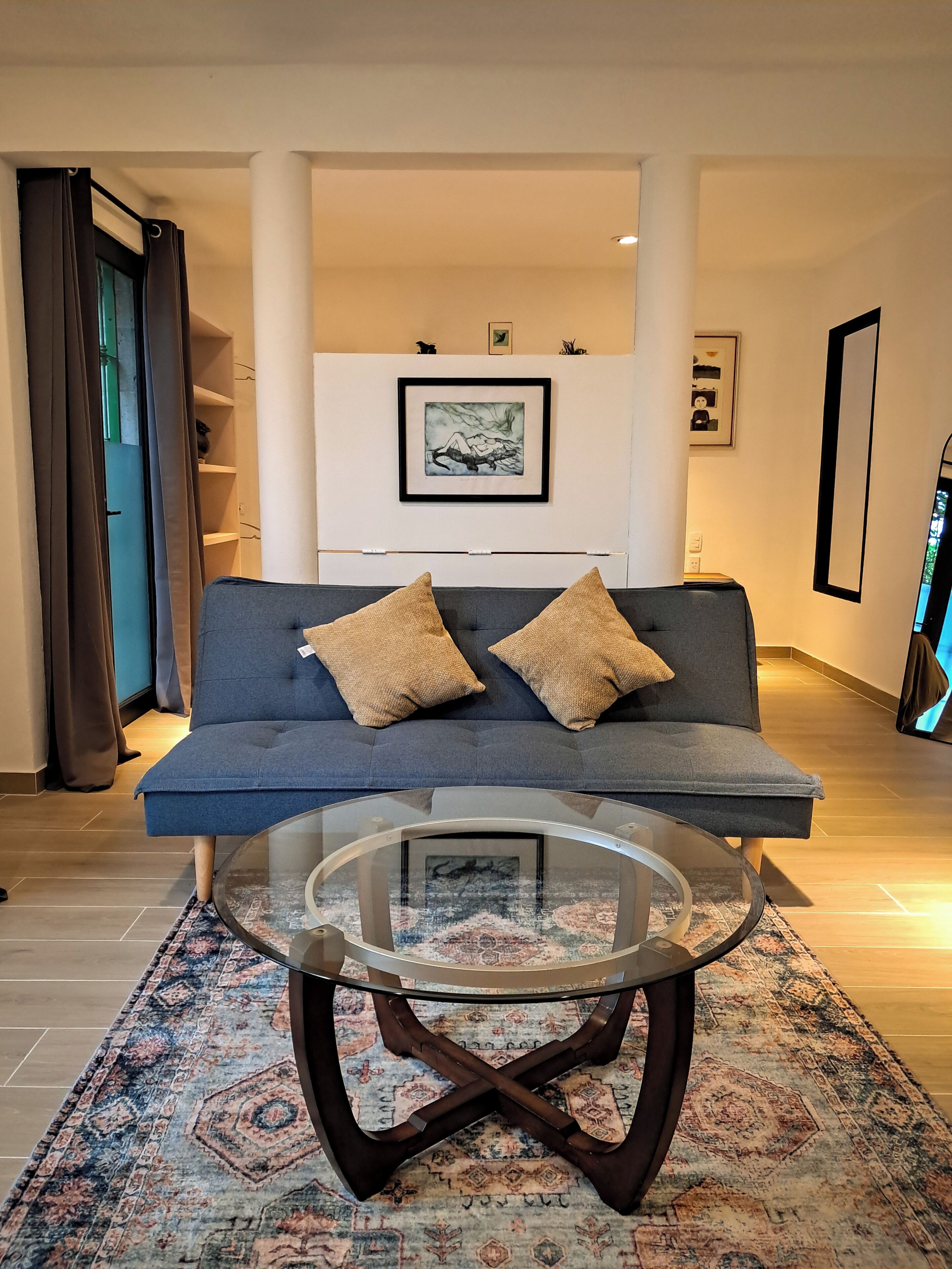 Una sala de estar moderna con un sofá azul, dos cojines marrones, una mesa de centro de vidrio y un tapete estampado.
