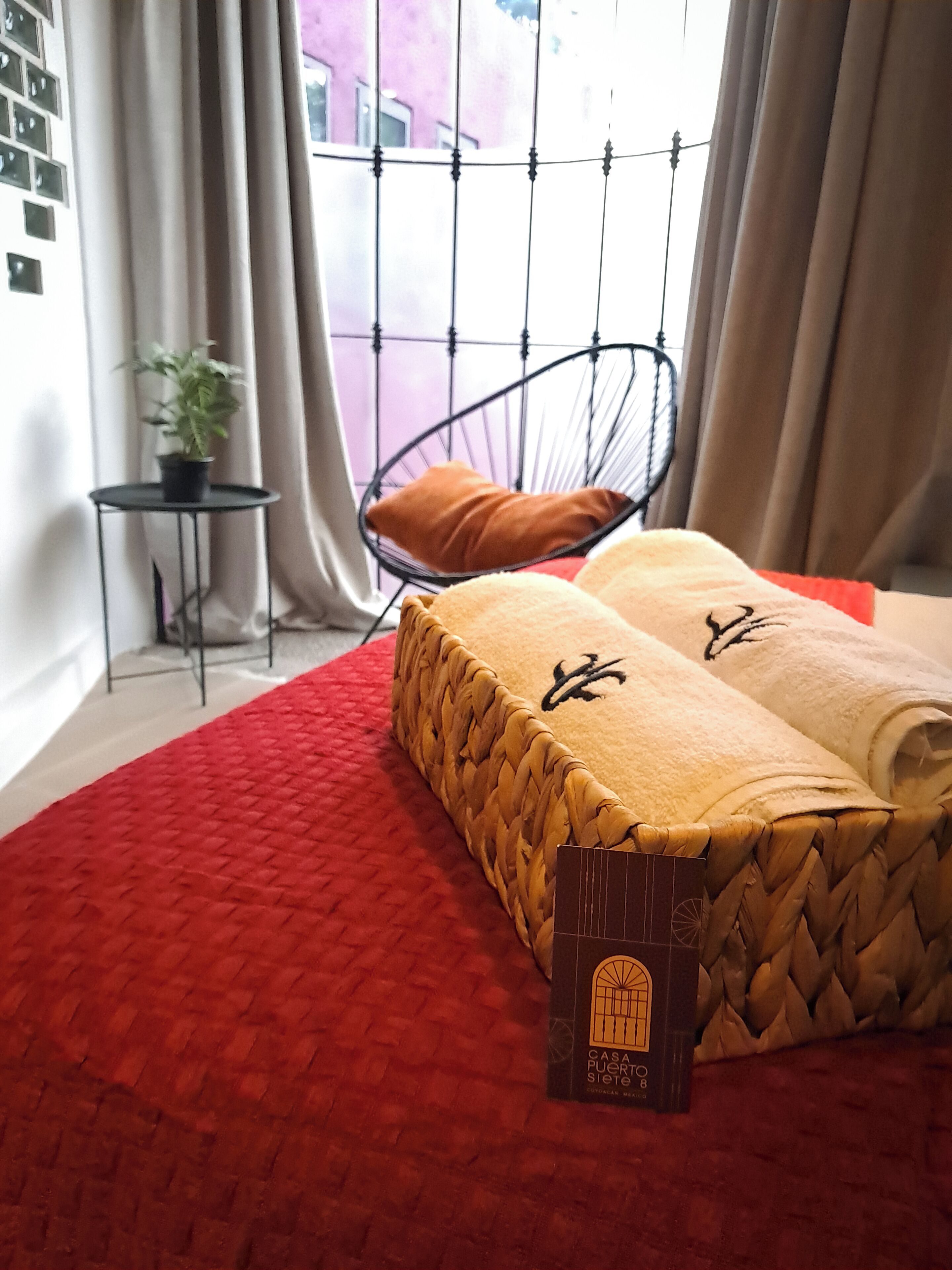 A bed with a red cover, a hanging chair, a small table with a plant, and a window with curtains.