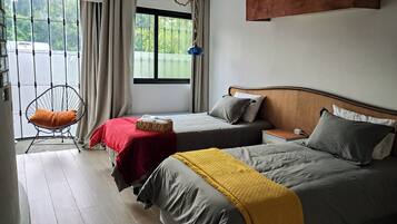 Quarto de hotel com duas camas, uma cadeira, janela com cortinas e vista para o verde lá fora.