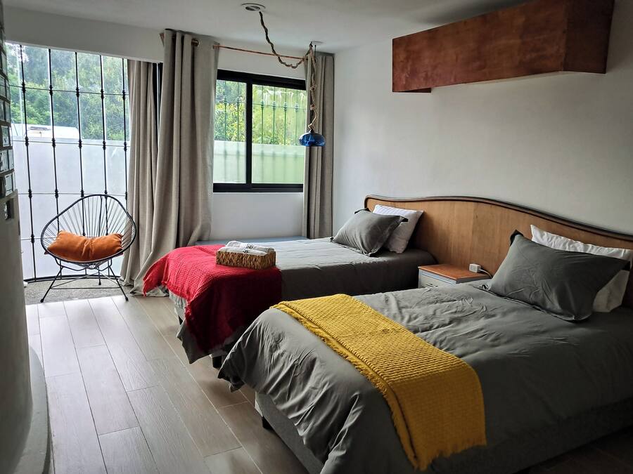 Quarto de hotel com duas camas, uma cadeira, janela com cortinas e vista para o verde lá fora.