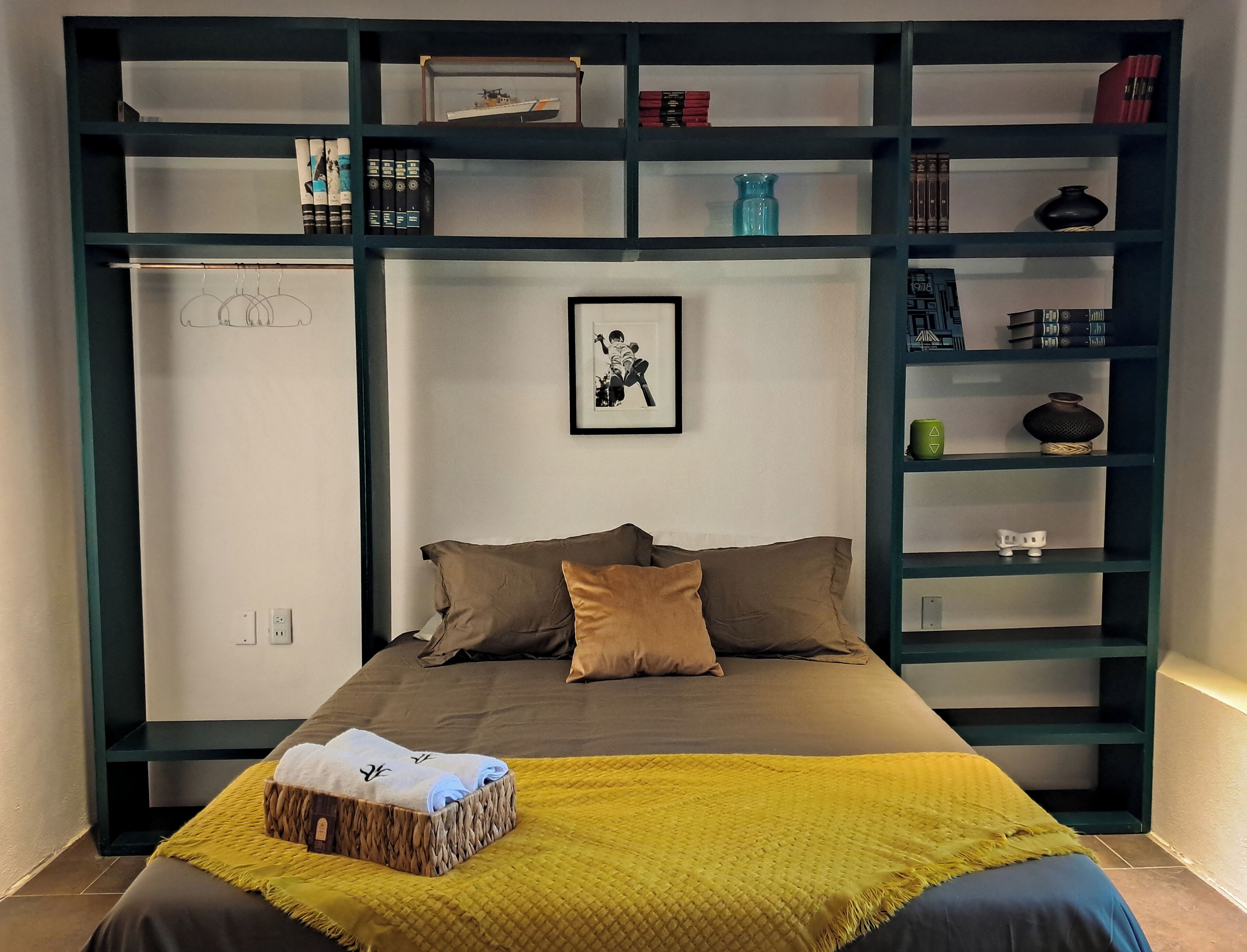 A bedroom with a bed, pillows, a yellow blanket, a framed picture, and a bookshelf with various items.