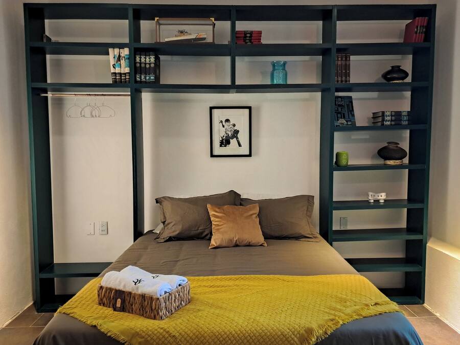 A bedroom with a bed, pillows, a yellow blanket, a framed picture, and a bookshelf with various items.