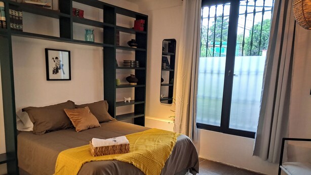 A bedroom with a bed, a bedside table, a window, and a shelf with various items. - Casa Puerto Siete8 (Mexico City)