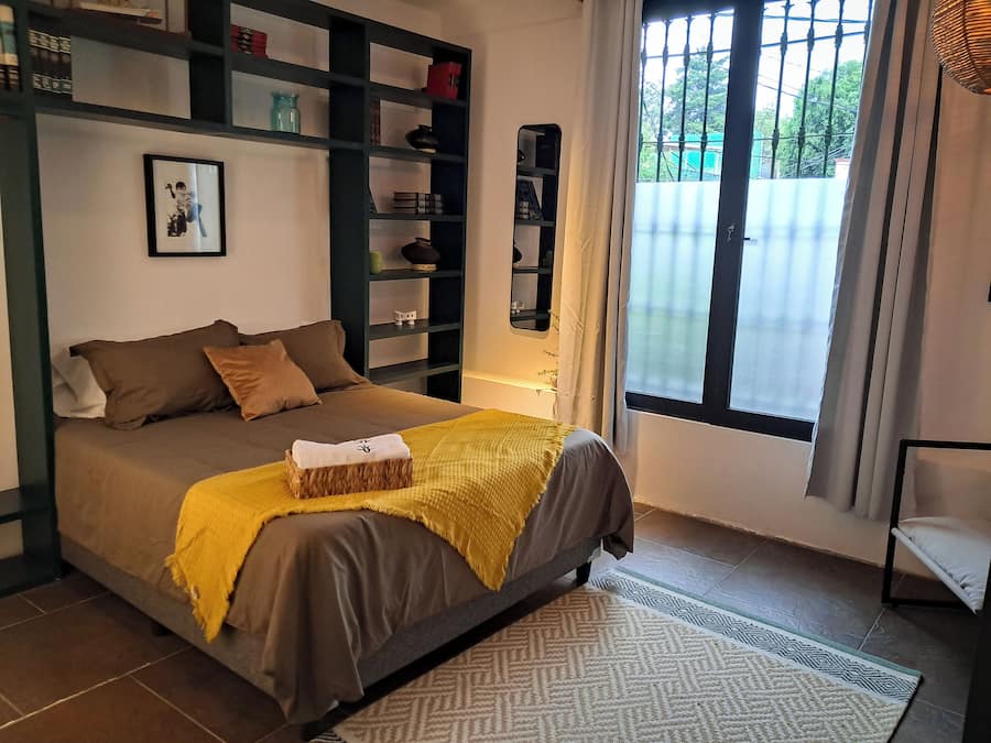 A bedroom with a bed, a bedside table, a window, and a shelf with various items.