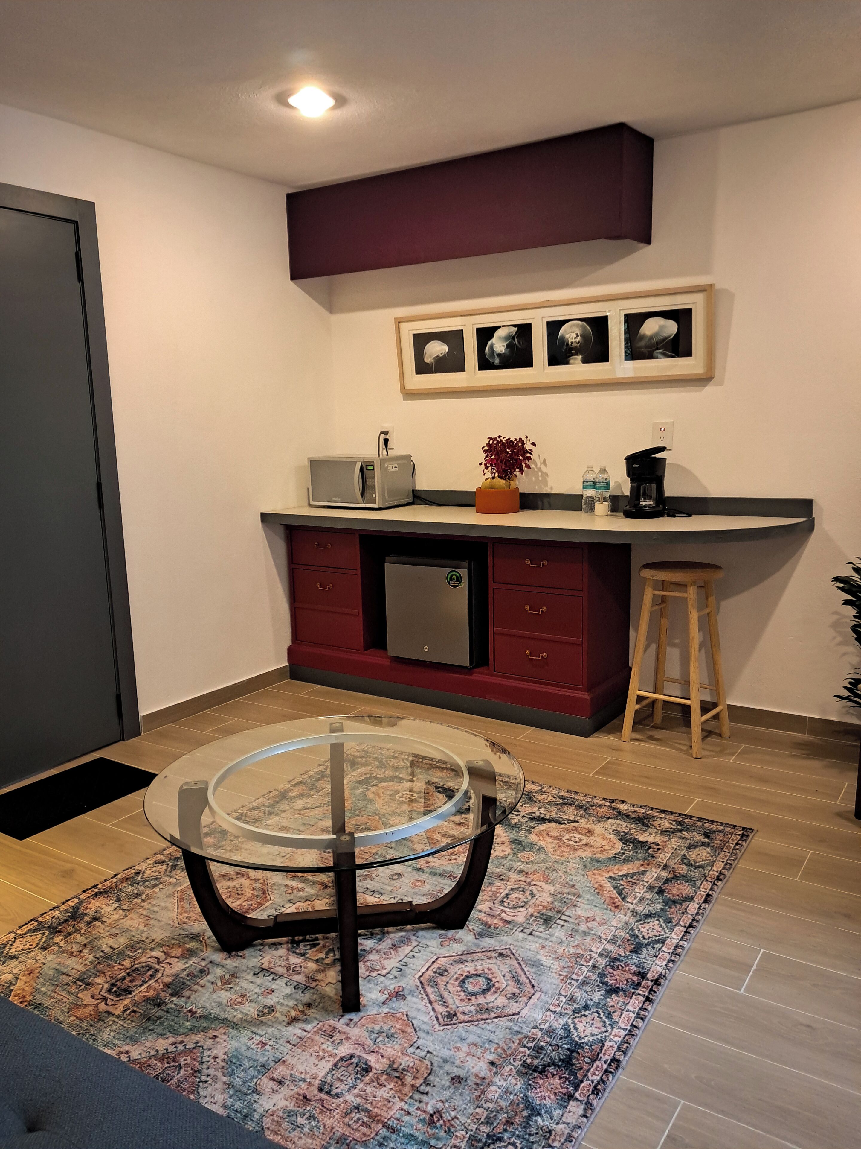 A compact kitchen with a microwave, toaster, and coffee maker, a small stool, and a glass table with a rug.