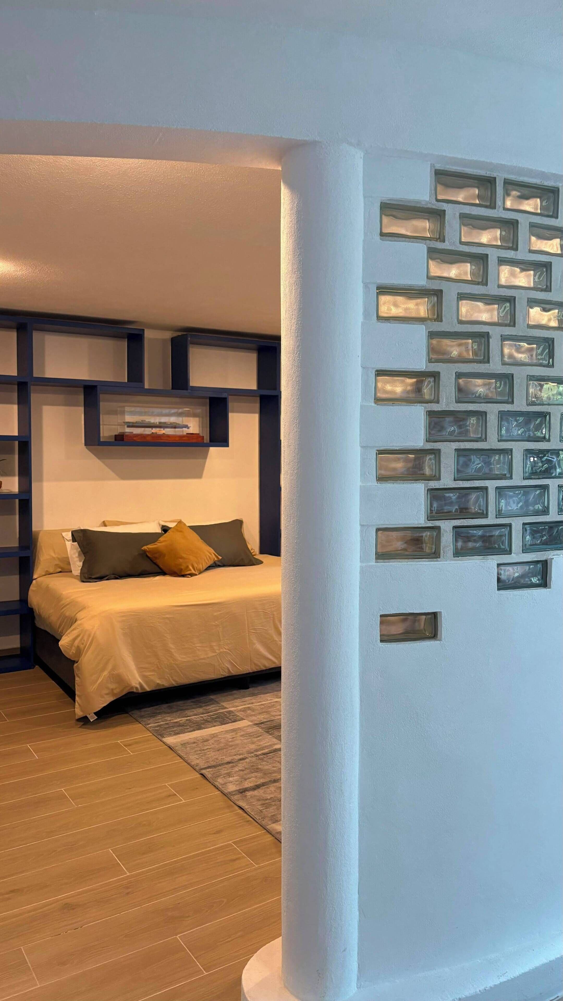 Quarto com cama, mesa de cabeceira, prateleira com uma imagem e painéis de vidro decorativos na parede.