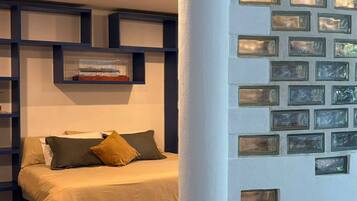 Quarto com cama, mesa de cabeceira, prateleira com uma imagem e painéis de vidro decorativos na parede.