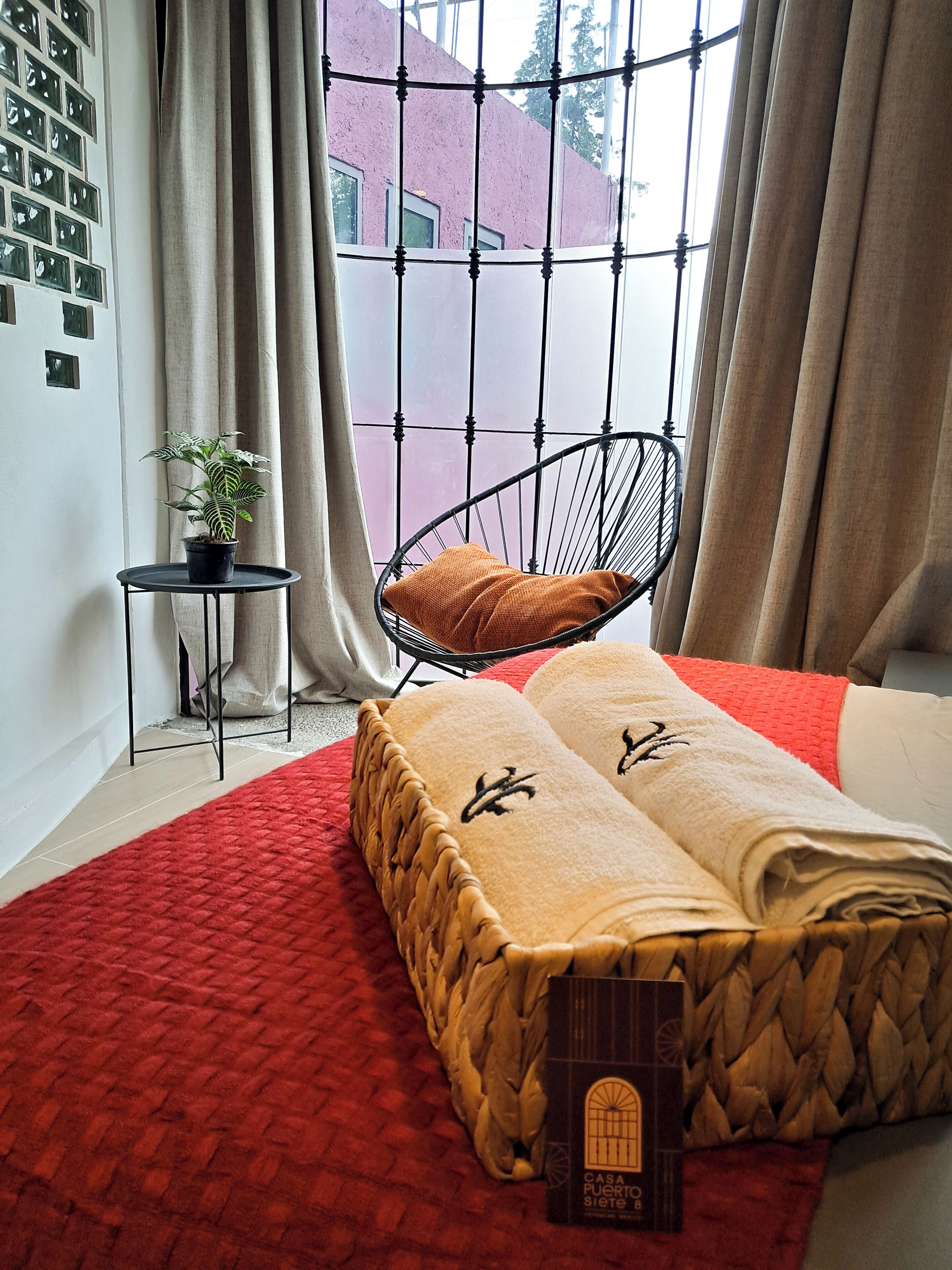A room with a bed, a chair, a small table with a plant, and a window with curtains.