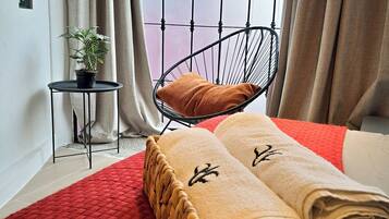 A room with a bed, a chair, a small table with a plant, and a window with curtains.