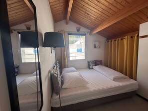 A bedroom with two beds, wooden ceiling beams, a window with curtains, and a mirror. - Apartment 200m from Beach, Pets & Parking Allowed (La Tranche-sur-Mer)