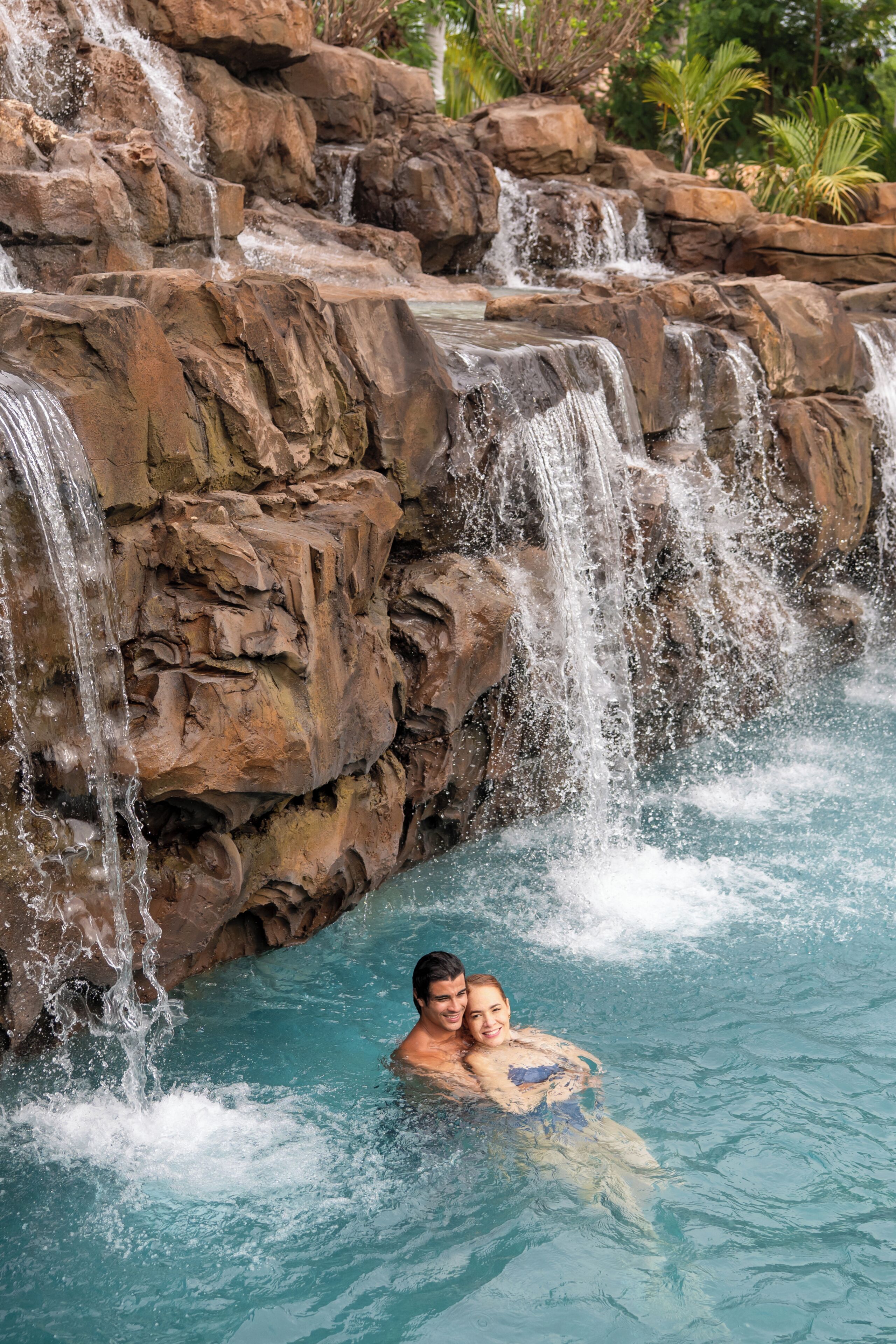 A man and a woman in a pool with a waterfall feature.