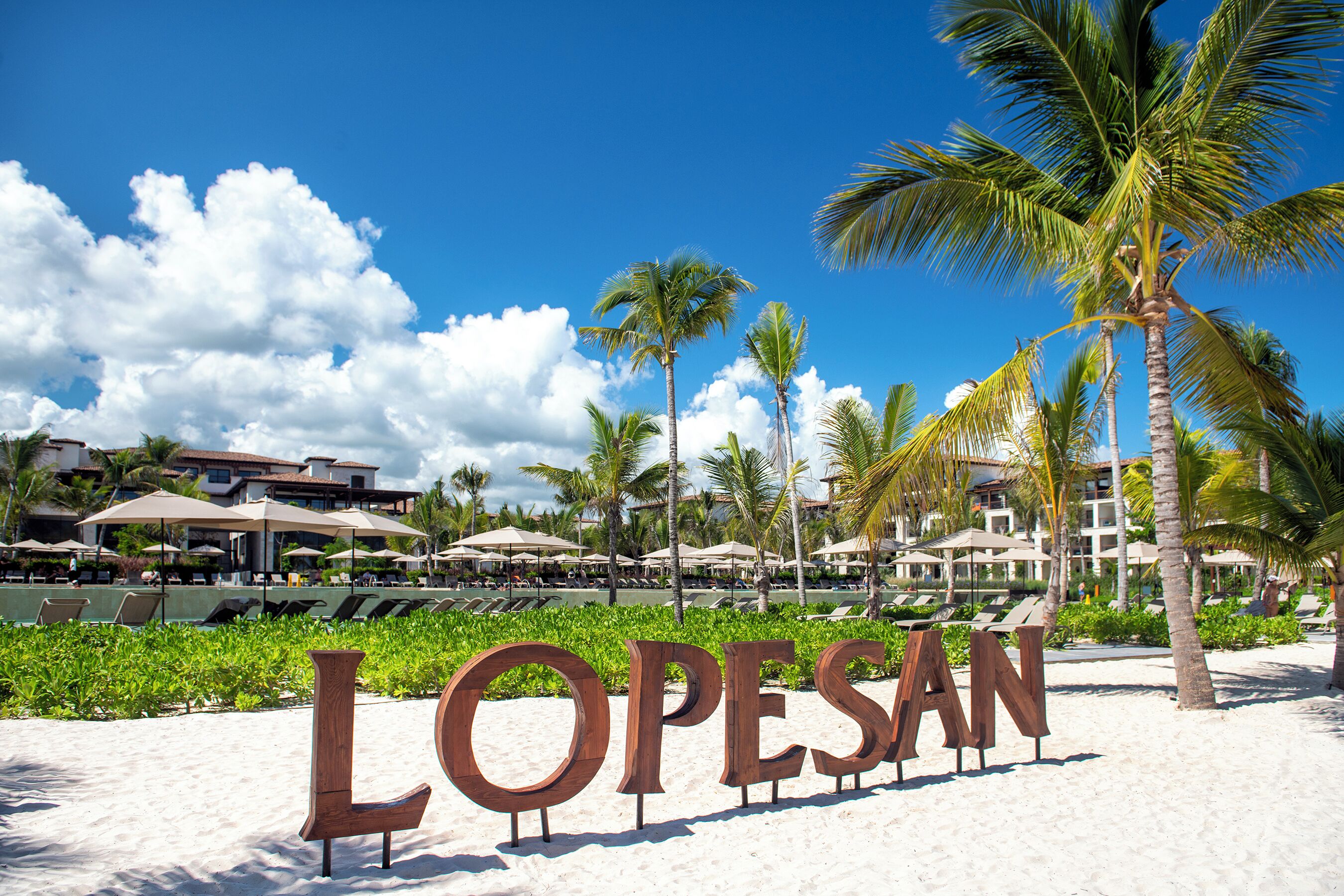 A beach with the word "LOPESAN" in the sand, palm trees, and a resort in the background.
