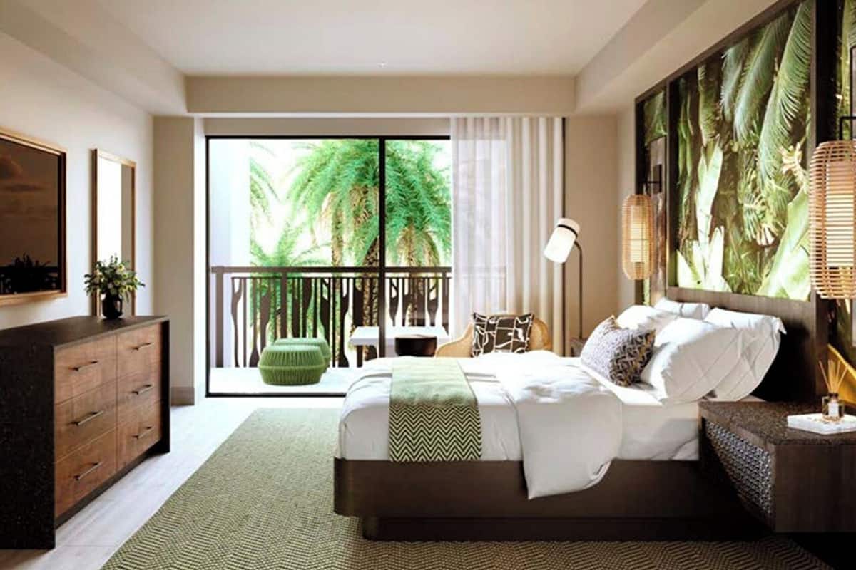 Une chambre moderne avec un grand lit, une vue sur les palmiers à travers une porte coulissante, un tapis à motifs verts et une commode en bois.