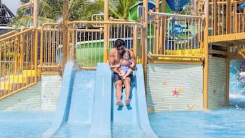A water slide with a man and a child at the top, surrounded by metal railings and palm trees.