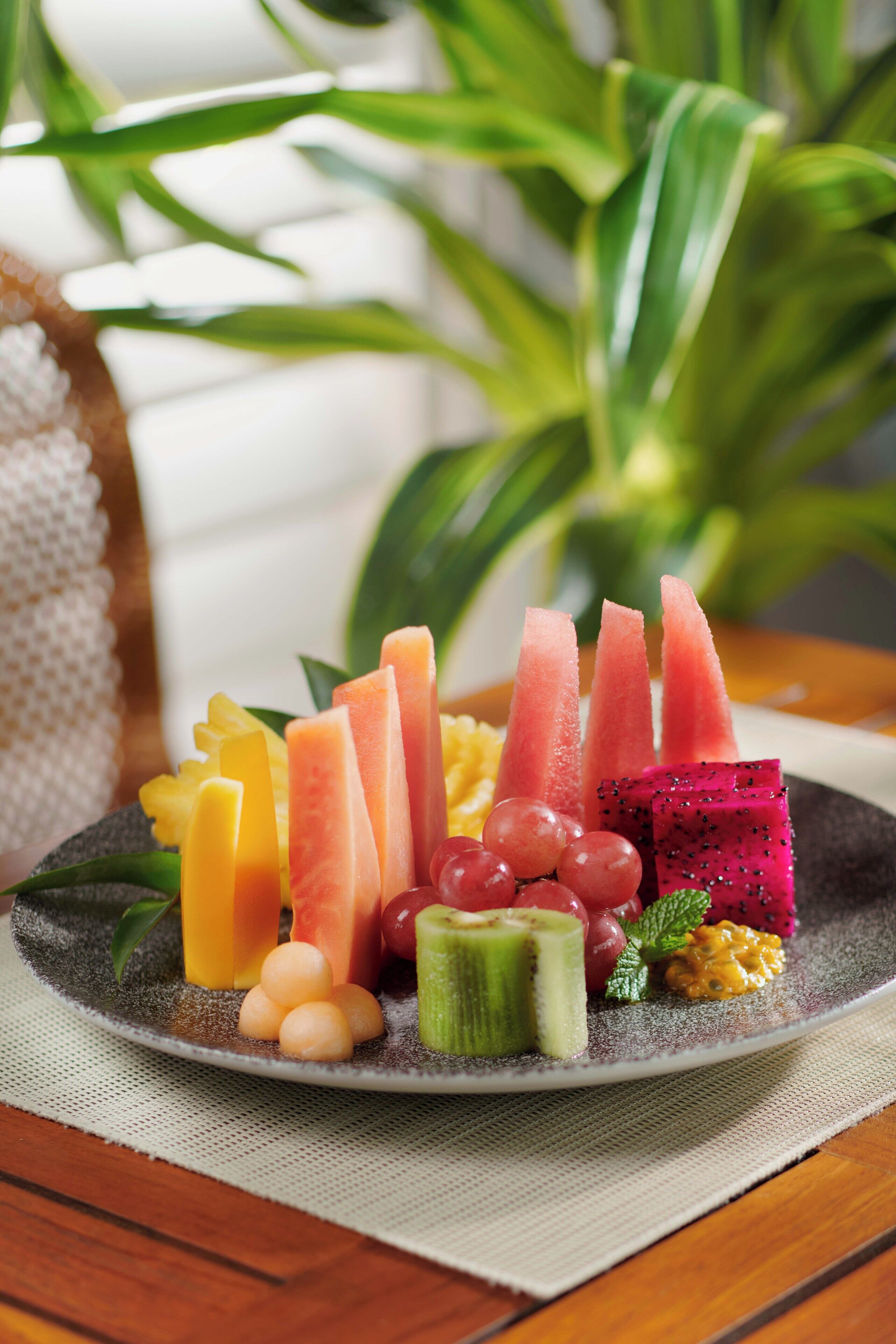 A plate of assorted fruits including watermelon, grapes, and dragon fruit, placed on a textured mat.
