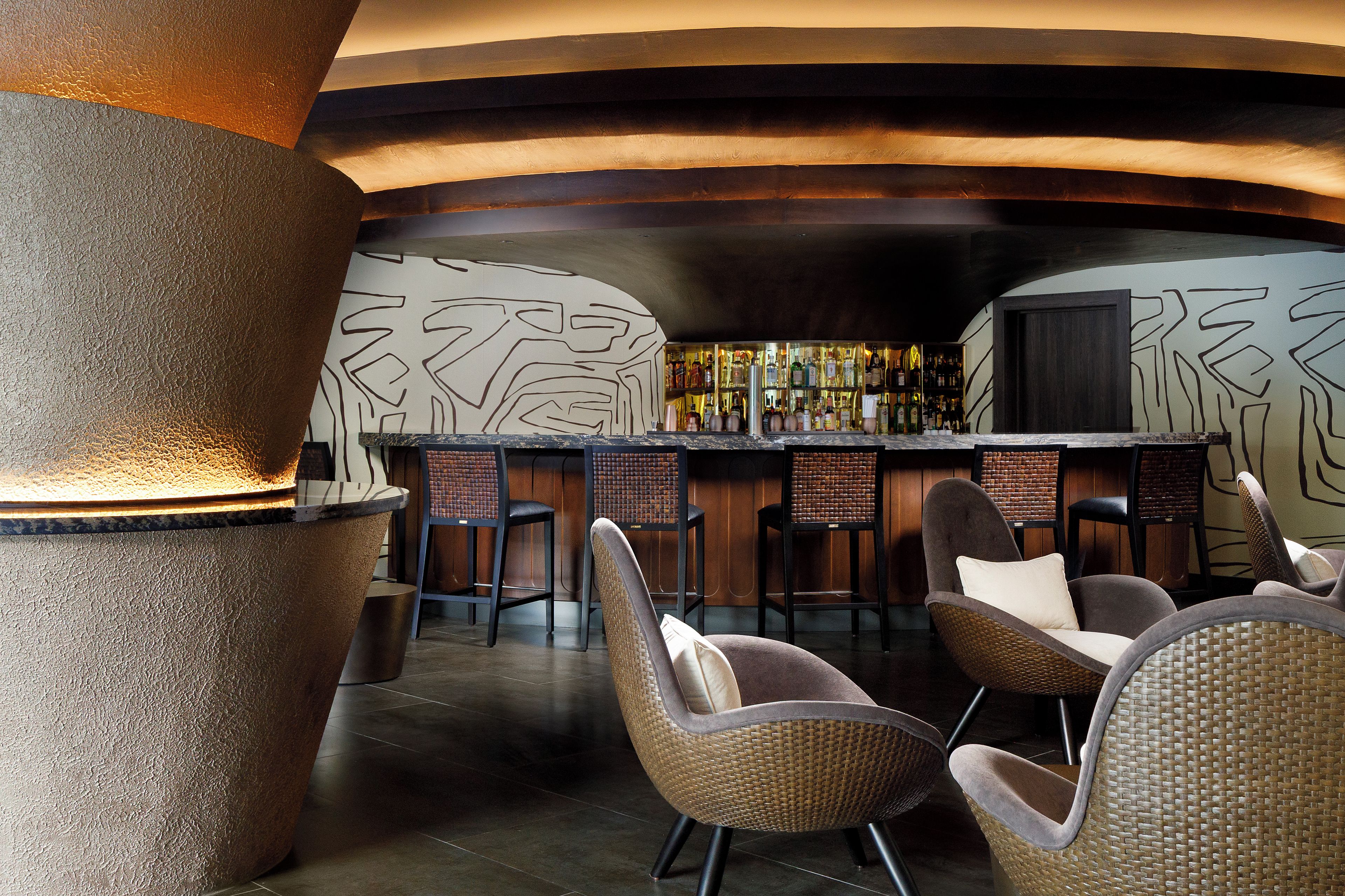 Un espace bar avec des chaises en osier, un pilier en pierre et un mur orné de motifs abstraits.