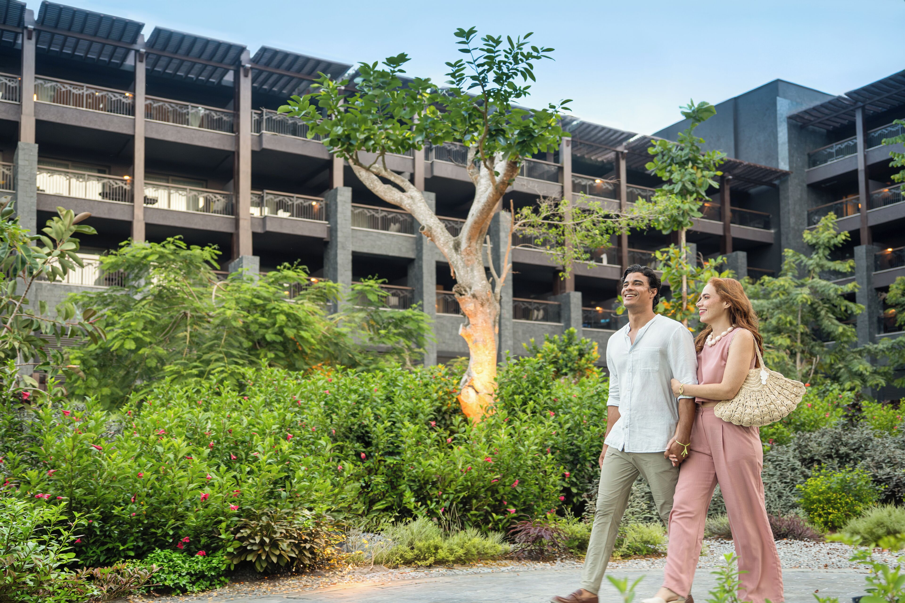 A couple walking in a courtyard with a tree, surrounded by greenery and a building with multiple balconies.