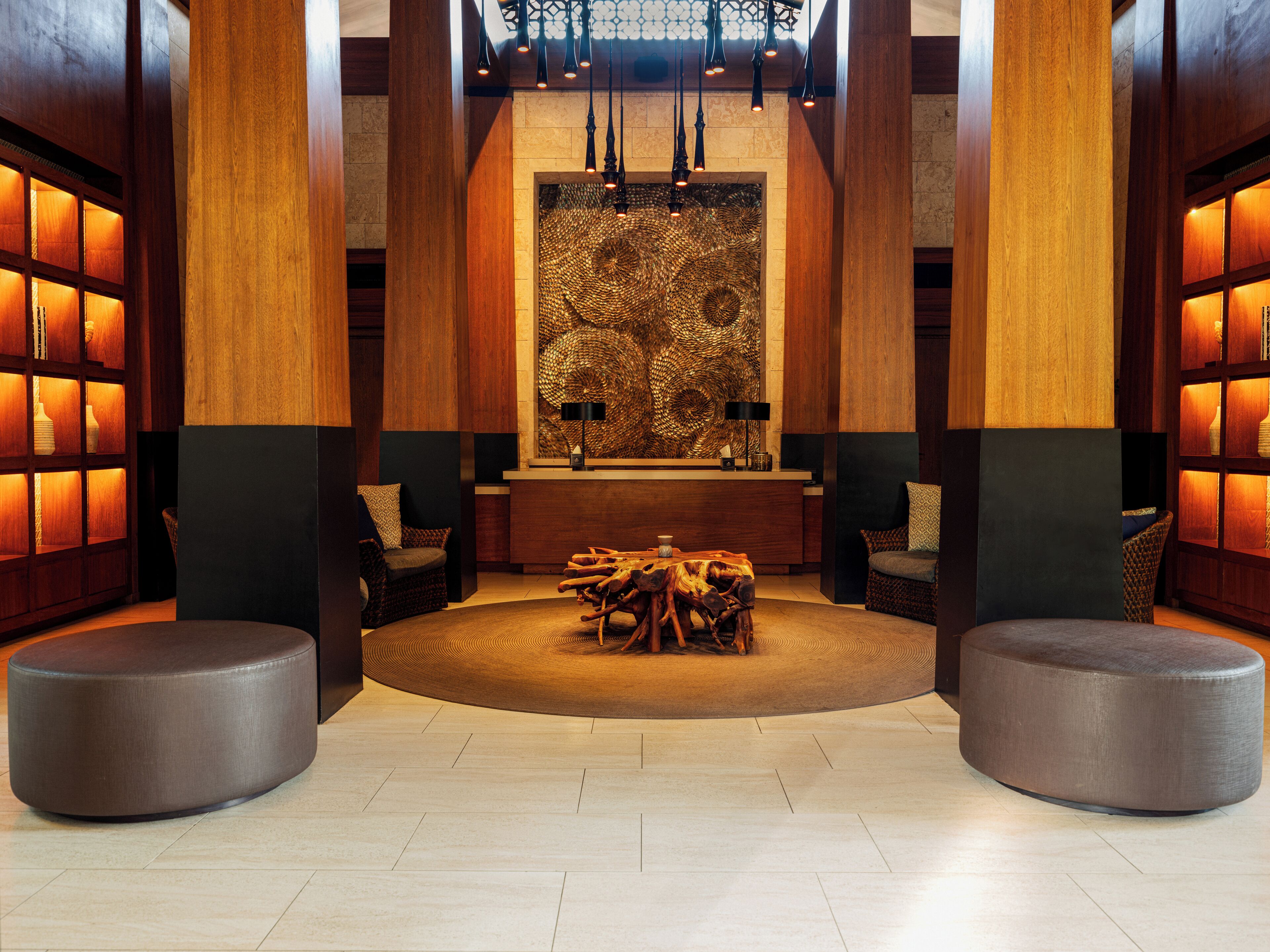 A lobby with a central reception desk, two seating areas with ottomans, a patterned wall, and wooden paneling.