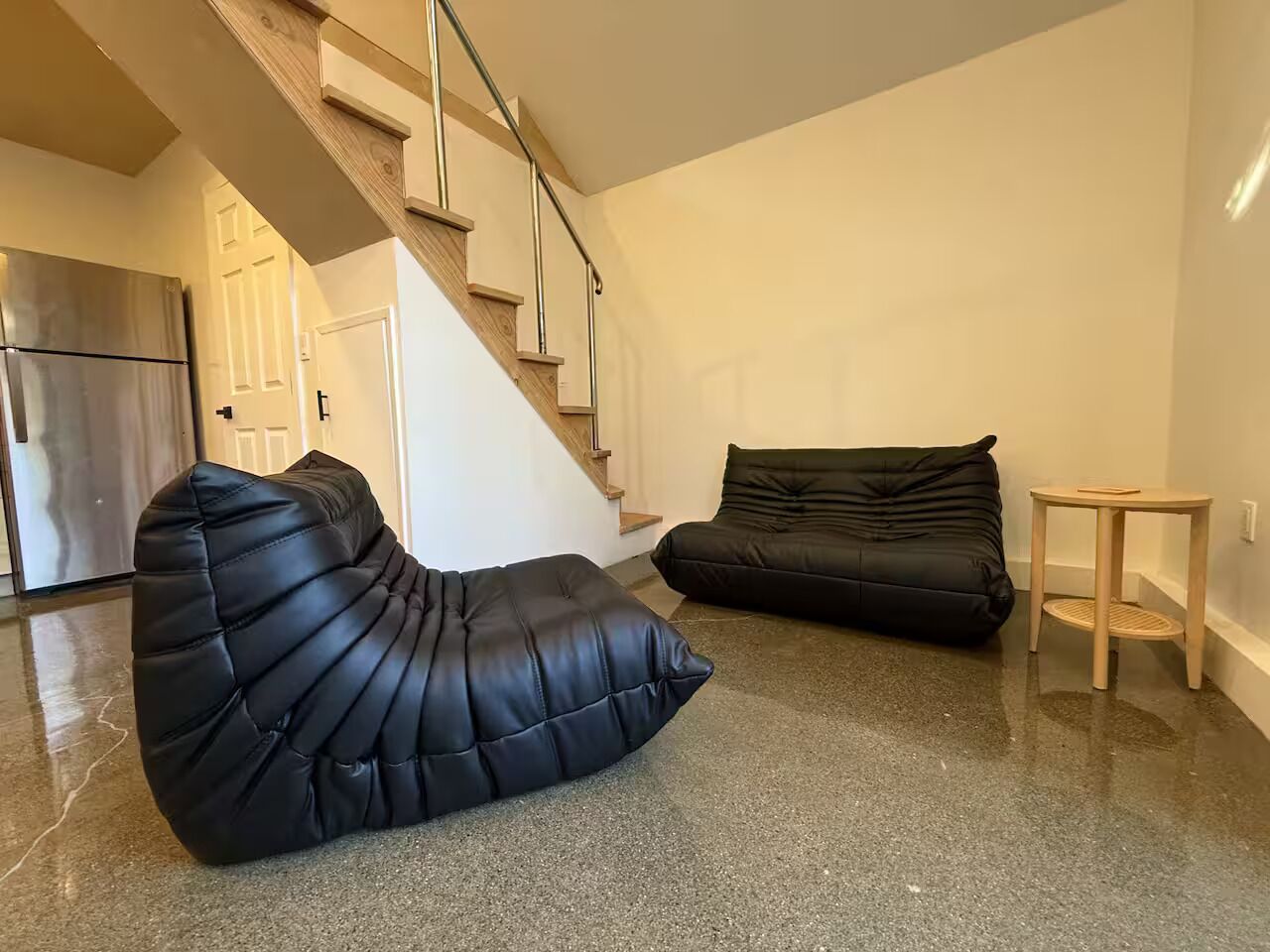A modern living room with a black pouf, a black sofa, a wooden coffee table, and a staircase leading to another level.