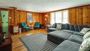 Ein gemütliches Wohnzimmer mit Holzwänden, einem Ecksofa, türkisfarbenen Sesseln und einem gemusterten Teppich.