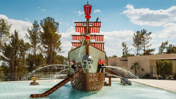Un toboggan aquatique sur le thème des pirates, avec une figure de crâne à l’avant, entouré d’arbres et sous un ciel dégagé.