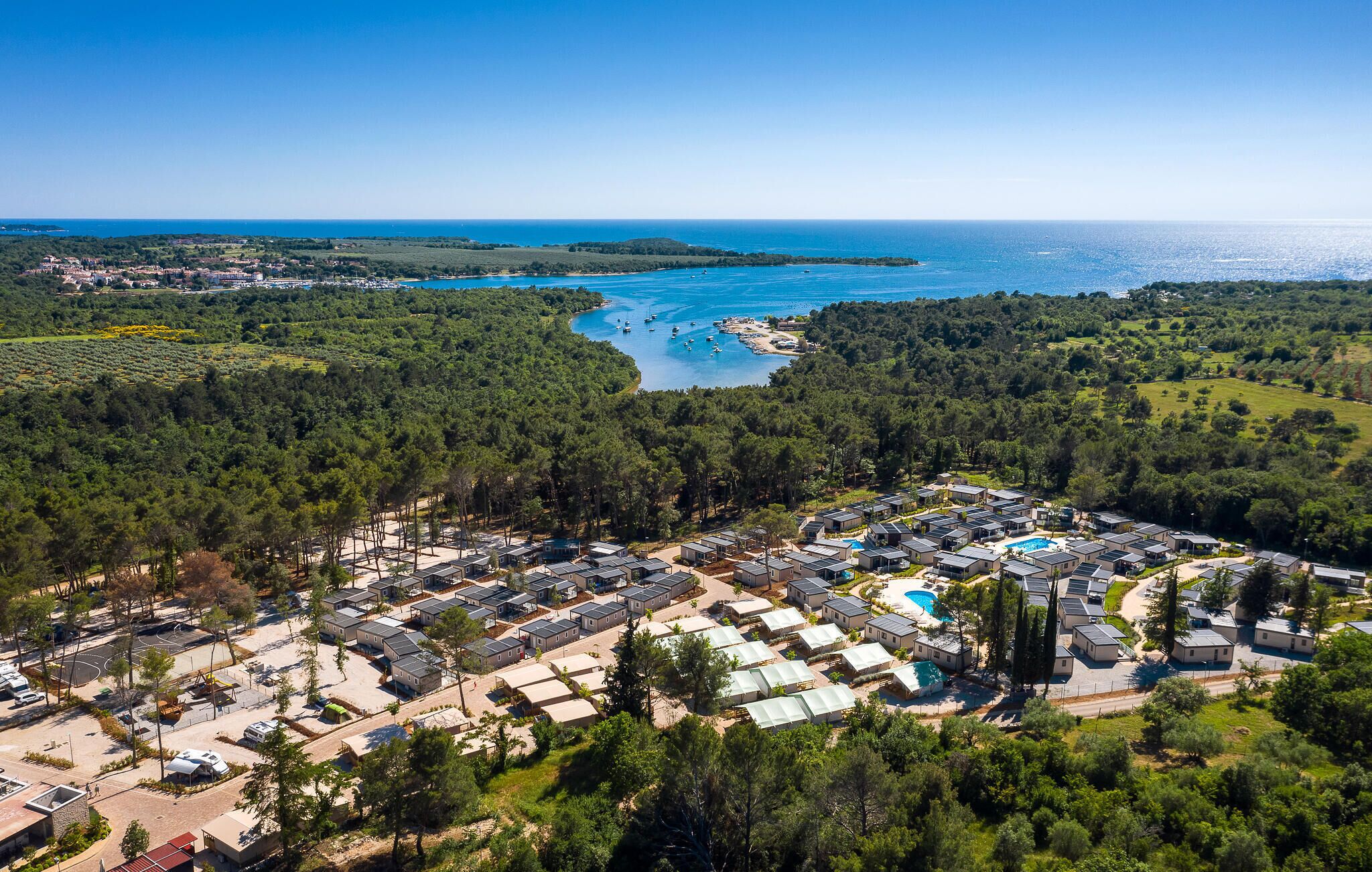 Vista aérea de un complejo turístico costero con múltiples alojamientos tipo bungalow, una piscina central y un puerto deportivo con barcos.