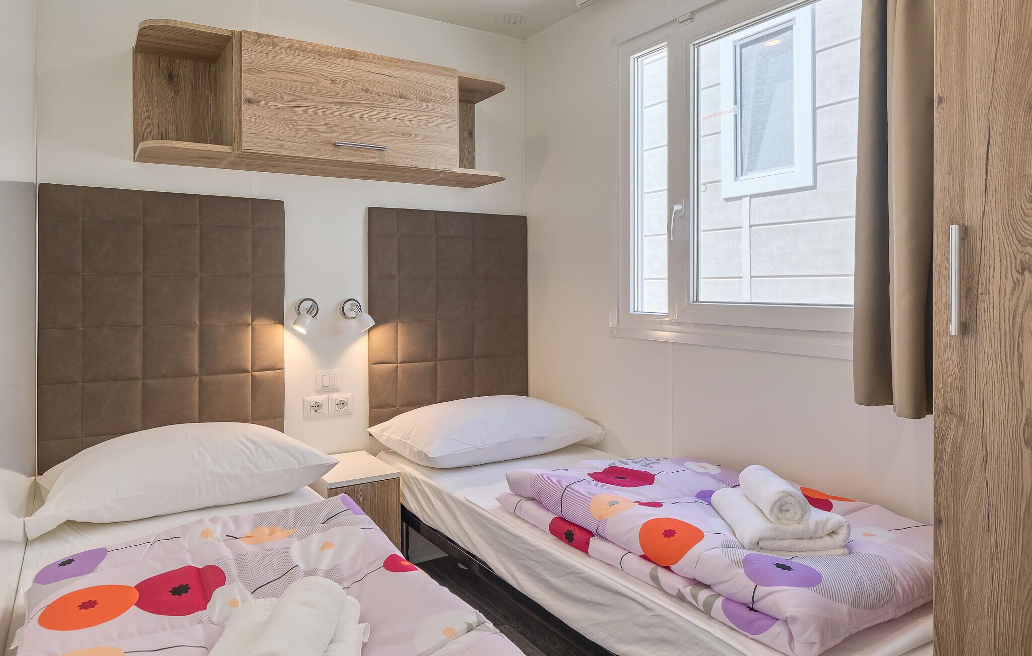 Habitación de hotel con dos camas, un estante de madera sobre ellas, una ventana con cortinas y una lámpara fijada a la pared.