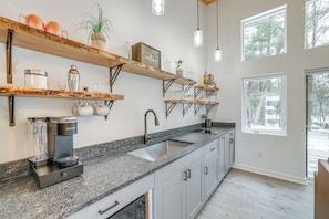 A kitchen with a granite countertop, a Keurig coffee maker, a sink, and multiple shelves with various items. - On-Site Trails & Trout Stream! NW Michigan Escape (Hersey)