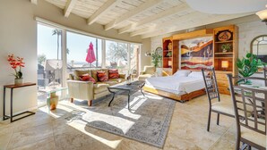 A spacious room with a bed, a sofa, a coffee table, and a dining area. There is a large window providing natural light and a view of palm trees. - Catalina Foothills Gem 8 Mi to Sabino Canyon (Tucson)