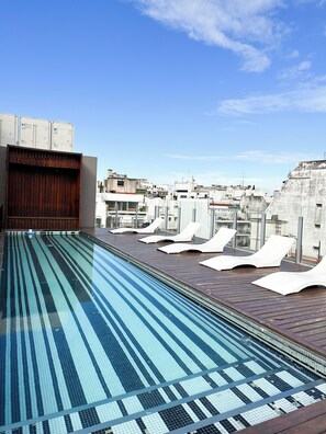 Una piscina en la azotea con cielo azul despejado, terraza de madera y sillones de descanso.